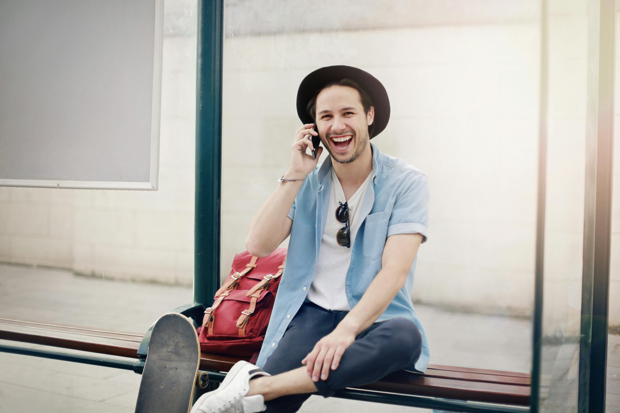 Smiling man wearing a hat and sunglasses sits on a bench at a bus stop, chatting on his phone.